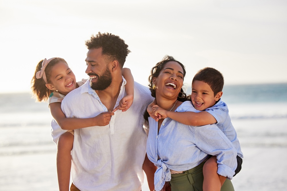 Happy family enjoying a fun day at the beach – Point Loma Family Dentistry Smiling parents carrying their young son and daughter on their backs while walking along the shoreline, representing healthy family smiles – Point Loma Family Dentistry