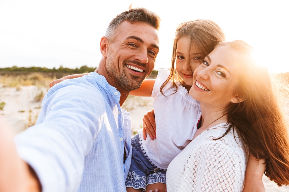 Smiling family taking a cheerful beach selfie at sunset – Point Loma Dentist Happy parents holding their daughter and posing for a sunny beachside selfie, showcasing bright smiles – Point Loma Dentist